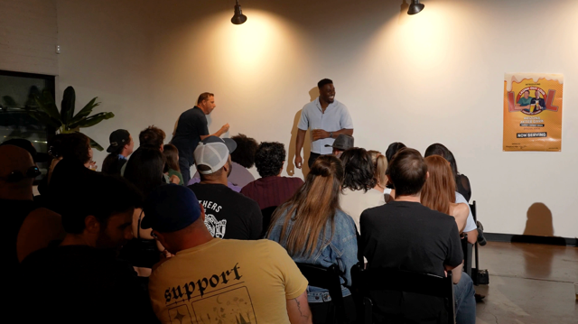 Two comedians on stage beside the LOL Beer poster in a packed room