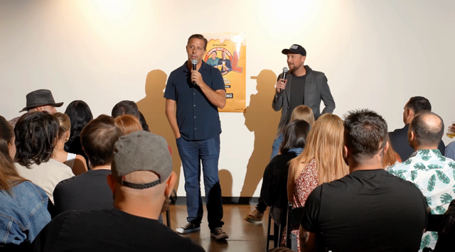 Host and comedian trading jokes on the mic in front of the show poster